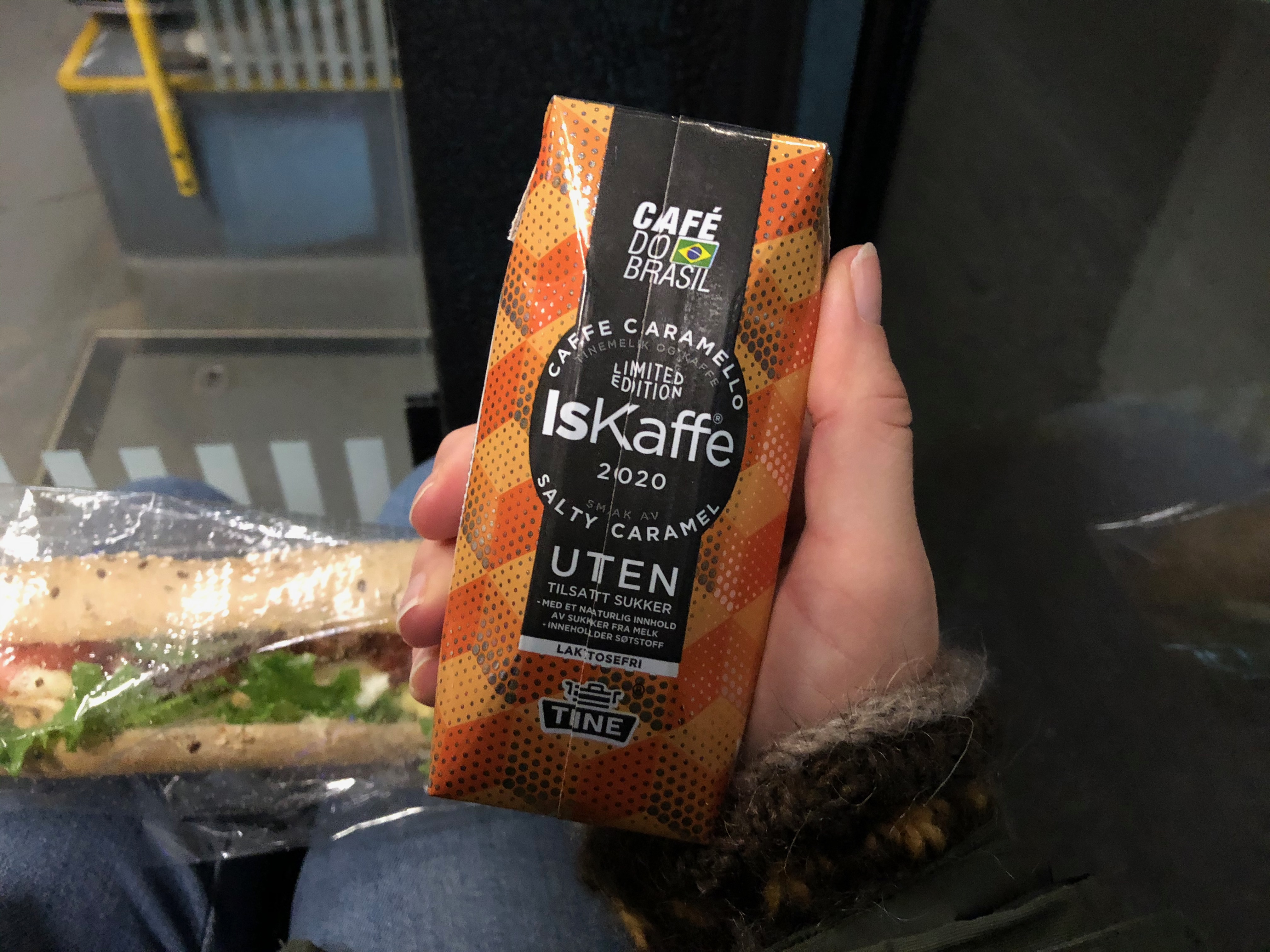 Meg som holder en boks med Tine IsKaffe Limited Edition 2020 CAFFE CARAMELLO Salty Caramel UTEN laktosefri. I bakgrunnen ligger en baguett.